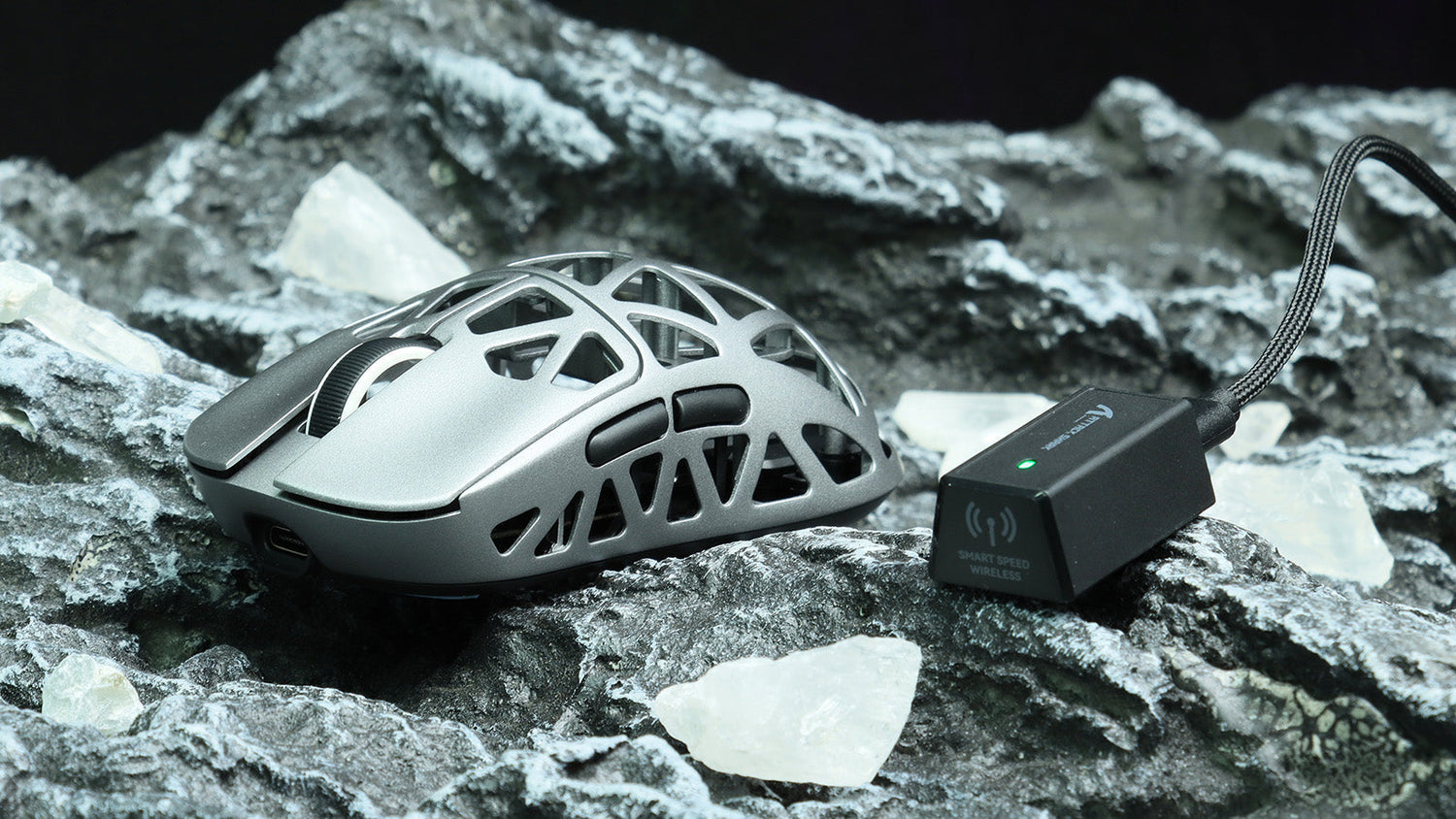 A silver lightweight gaming mouse with an open exoskeleton shell, resting on a rocky surface next to its "Smart Speed Wireless" dongle
